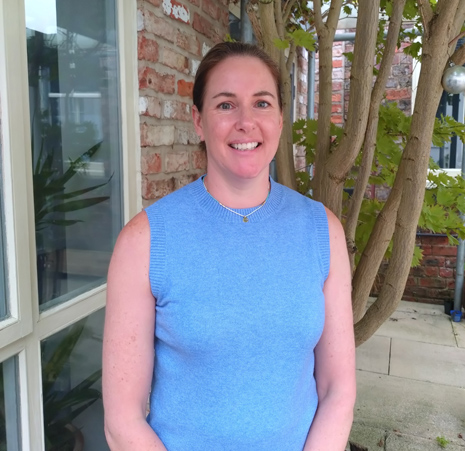 A woman with brown hair tied back in a sleeveless blue top stood by a tree.