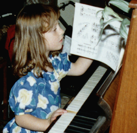 A young girl with brown hair and a blue and white flowery dress sits at a piano, turning the page of a book of sheet music.