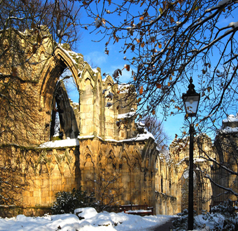 an abbey building made of yellow stone with an arched window frame in the snow, with a tree branches and a black old fashioned lamp post in front.