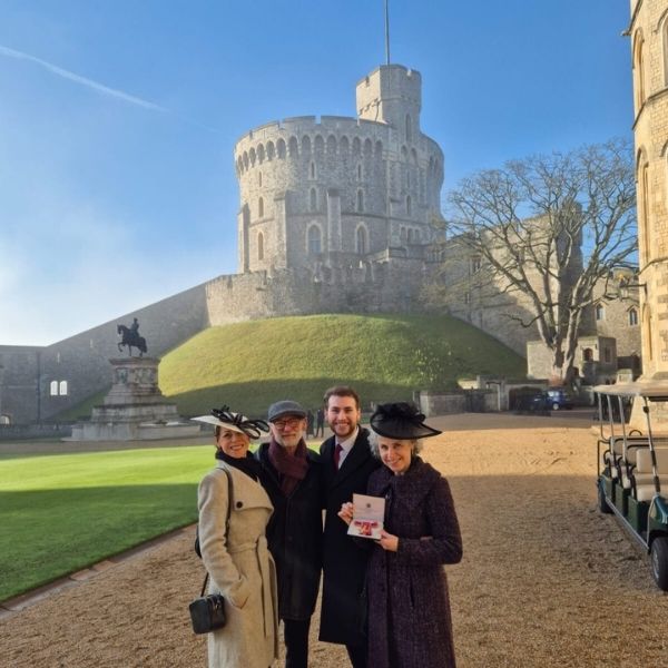 Windsor castle, a circular brick tower, against a blue sky with a family of four people stood in front, with Lesley in a black coat and hat holding the red ribbon of her OBE in a black box.