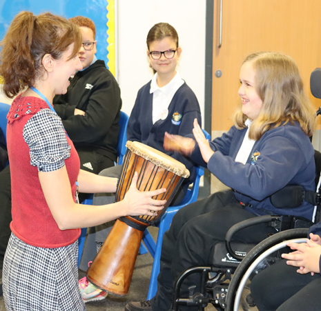 A woman with curly brown hair wearing a checked dress with a red tank top, is sharing a djembe drum with a young girl in a wheelchair, who has light brown hair, is smiling and playing the drum.