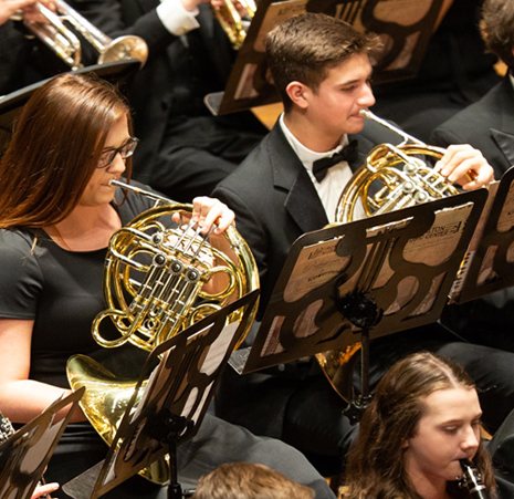 a young woman with brown hair and glasses wearing a black dress and a young man with dark hair, wearing a black suit with white shirt and bow tie, both playing the french horn.
