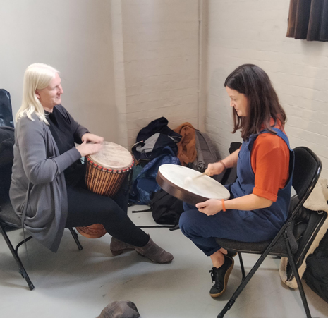 a women with white hair playing a djembe sat opposite a woman with dark hair wearing dungarees playing a round drum.
