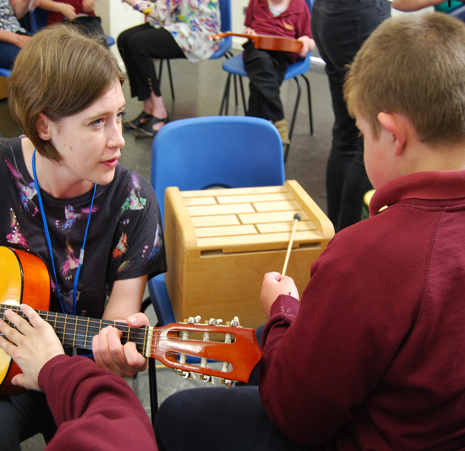 A woman with short brown hair is holding a guitar, a boy in a red jumper is sat playing a wooden musical box with a beater.